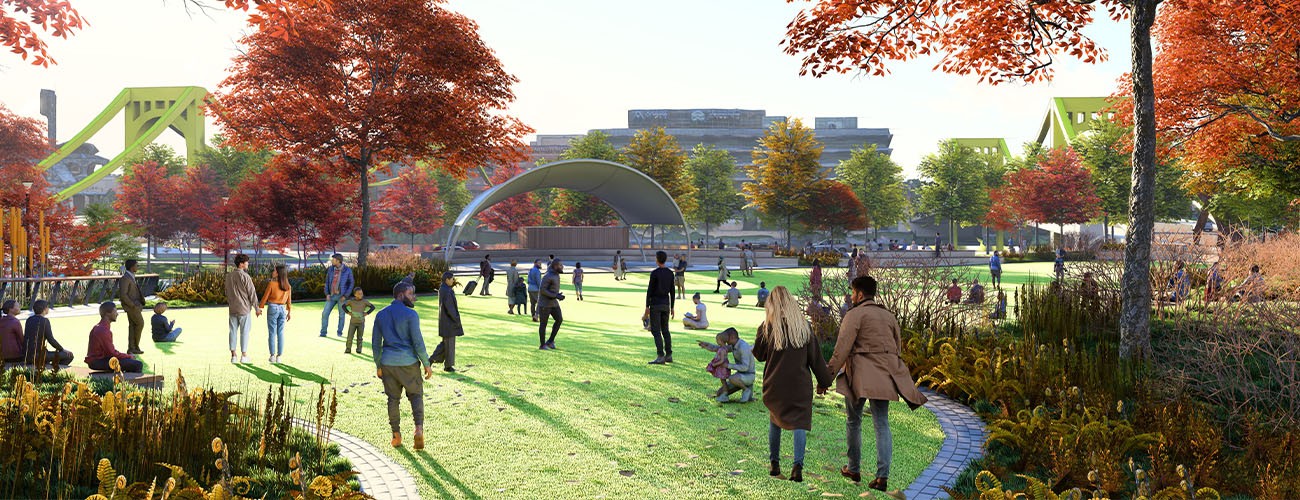 People walk and gather in a vibrant park with autumn trees, green grass, and a modern curved stage structure in the background; yellow bridges and city buildings are visible in the distance.